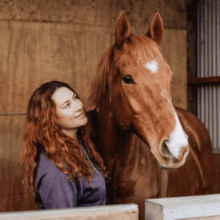 Young woman with long red hair and a chestnut horse in a stable.